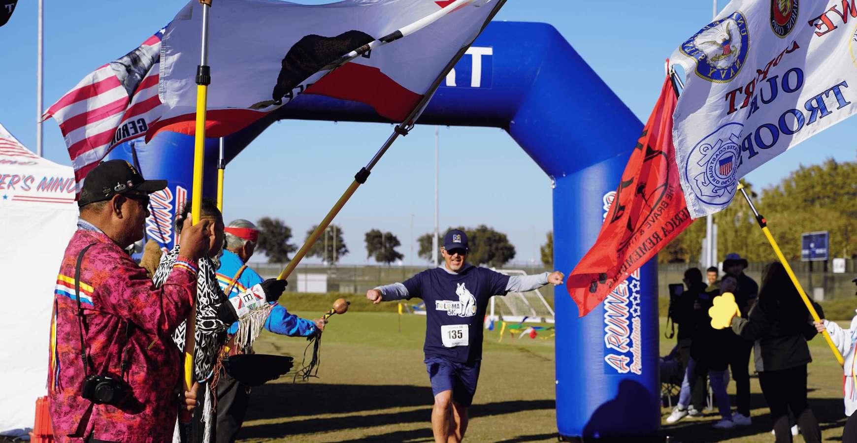 Alt text: A runner wearing a race bib crosses beneath a blue finish arch with arms outstretched as spectators on both sides hold military and California flags and cheer at the finish line of an outdoor 5K event.